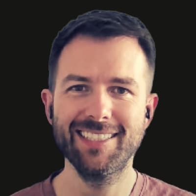 A person with short, neatly styled brown hair and trimmed facial hair, wearing a maroon crew-neck shirt. The individual is positioned against a solid black background, and is wearing small, dark in-ear headphones. The lighting is even, highlighting the hair and upper shoulders.