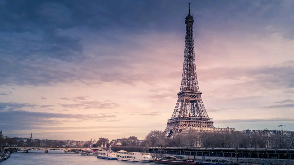 The Eiffel Tower in Paris at sunrise with the Seine River and boats in the foreground.