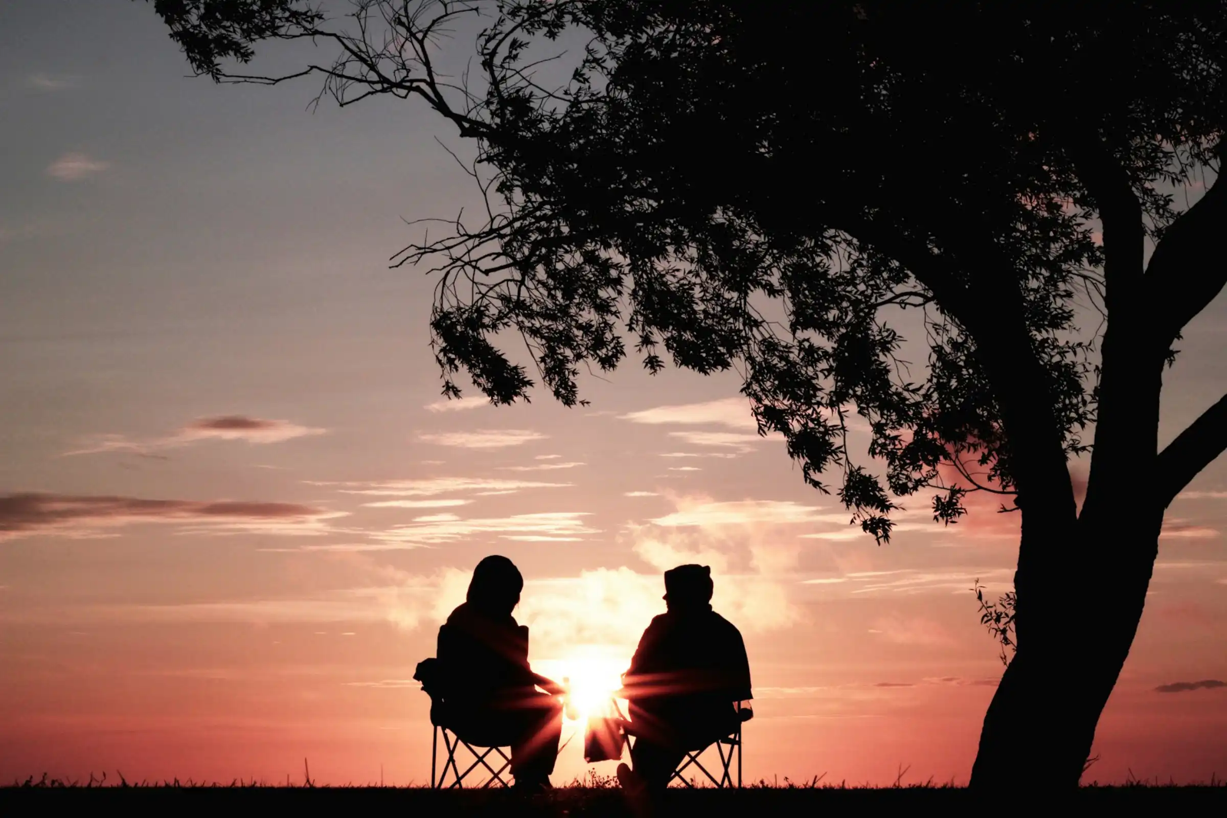 Two people sit in folding chairs under the shade of a large tree, silhouetted against a vivid sunset. The sky is filled with soft orange, pink, and purple hues, with thin clouds stretching across the horizon. The sun is low, shining brightly between them, casting a warm glow and sun rays outward. The scene feels calm and peaceful, suggesting a quiet moment of conversation or companionship in nature