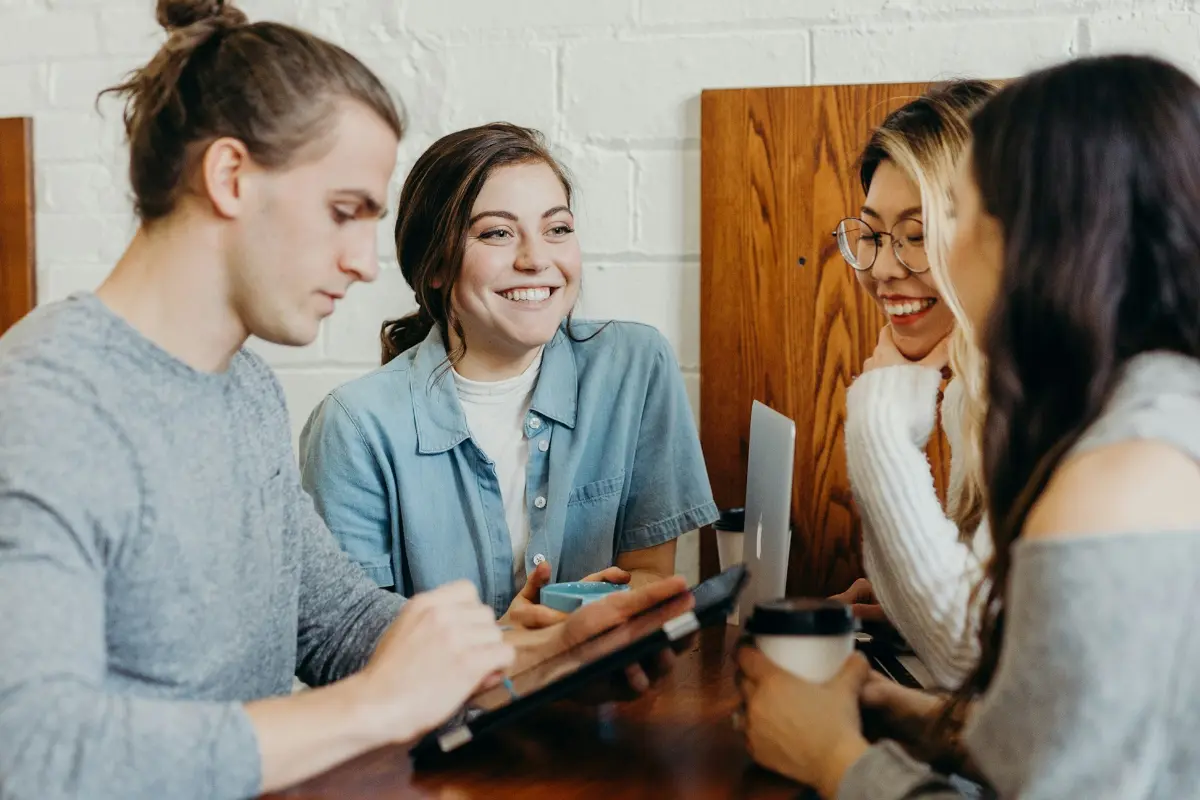 Four young adults sit together at a wooden table in a casual setting, possibly a café or study space. They are engaged in conversation and appear to be working collaboratively. One person is holding a tablet, another has a laptop in front of them, and two are holding coffee cups. Everyone is smiling or laughing, creating a warm, friendly, and collaborative atmosphere. The background includes a white brick wall and wooden panels, adding to the cozy, informal vibe.